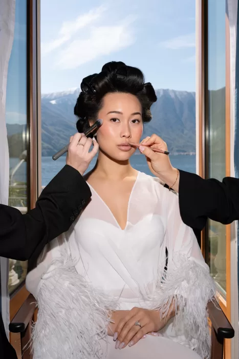 Bride having makeup applied by the window, with Lake Como softly visible outside.