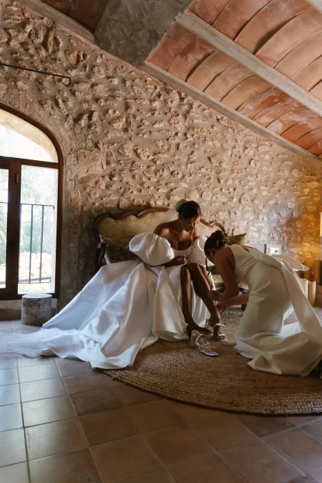Bride sitting on a bed in a rustic Mallorca finca room while a bridesmaid helps fasten her wedding shoes