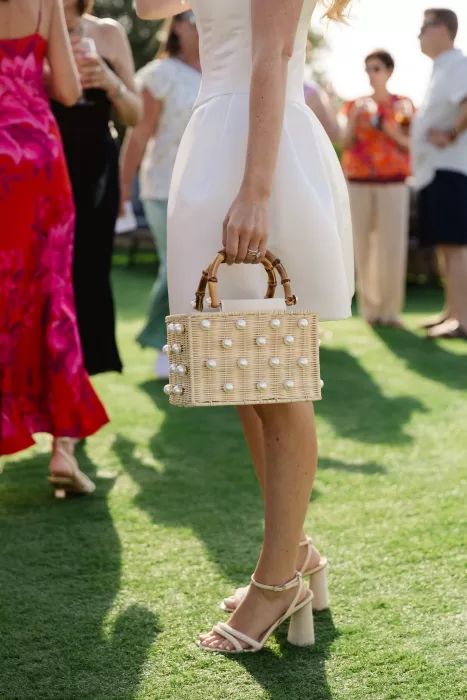 Bride holding a pearl-detailed wicker bag during a sunny outdoor drinks reception.
