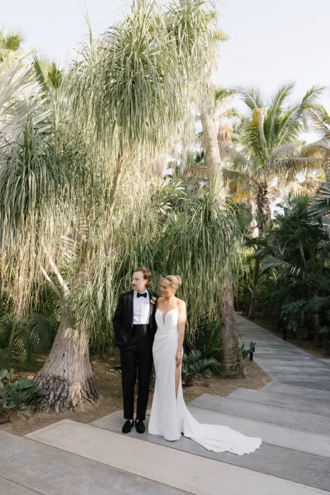 Bride and groom standing beneath sculptural palms along a quiet garden path.