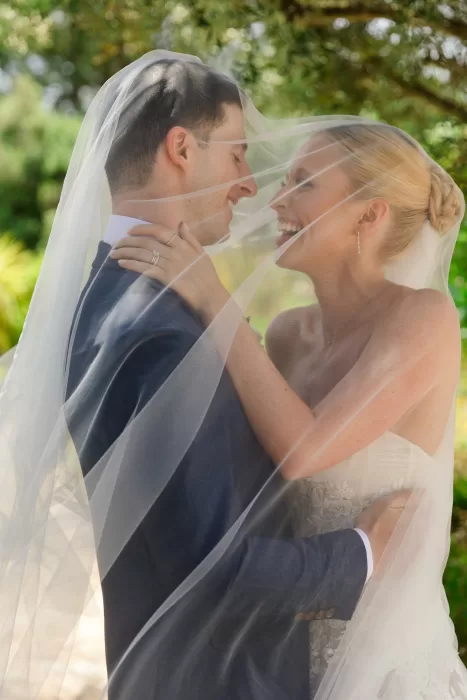Bride and groom laughing together beneath a veil in a lush garden setting.