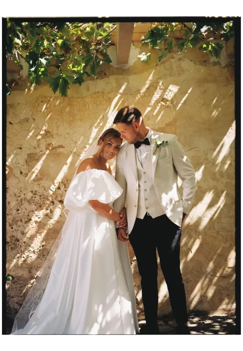 Bride and groom standing beneath vines, dappled sunlight casting soft shadows on rustic wall