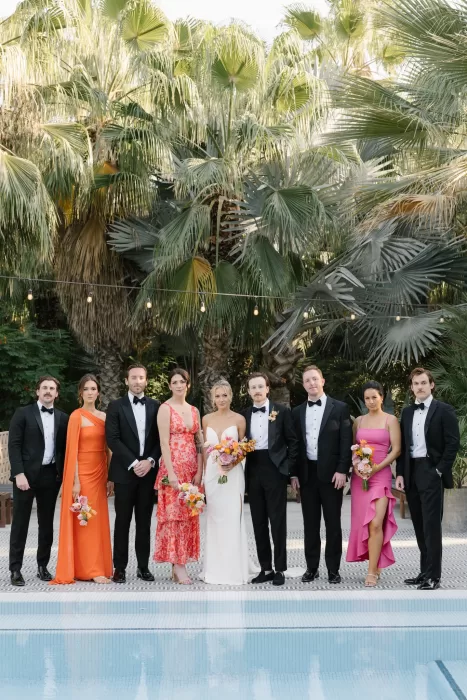Wedding party gathered poolside, with bright dresses contrasting black tuxedos and palms.