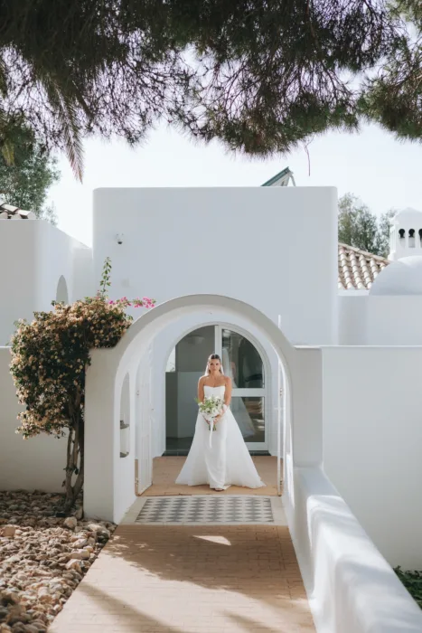 The bride stands beneath a white arched entrance holding a soft green and white bouquet.