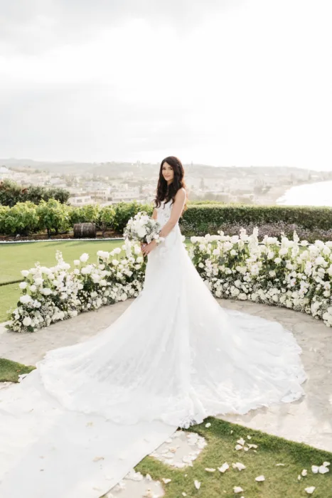 Bride in a lace gown stands among white flowers overlooking the bright sea.