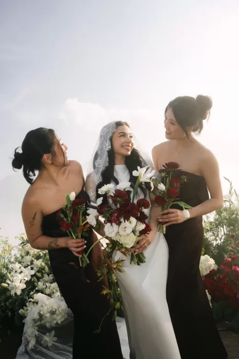 The bride laughing with her bridesmaids, surrounded by dark florals and high-altitude clouds.