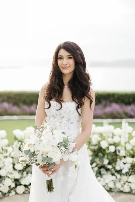 Bride holds a white rose and orchid bouquet against a soft coastal garden backdrop.
