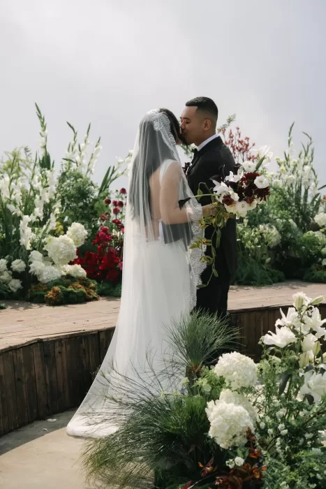 The couple kiss among white and burgundy flowers on a cloud-wrapped ceremony platform.