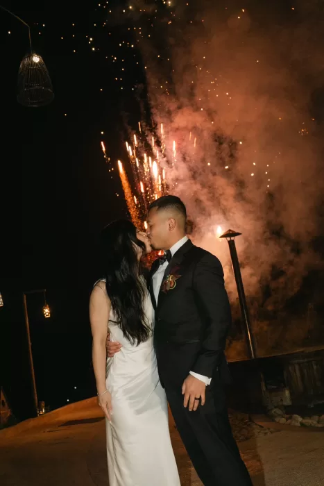 The couple kiss beneath glowing fireworks after dark, surrounded by smoke and warm light.