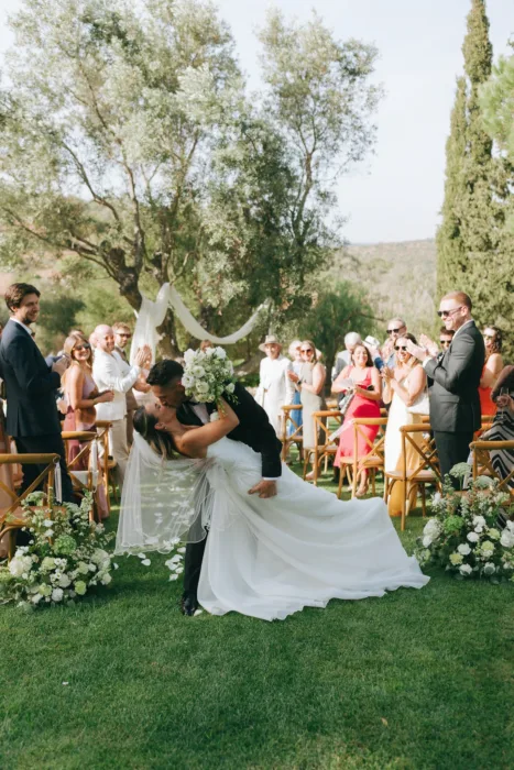 The couple share a celebratory ceremony kiss beneath olive trees as guests applaud.