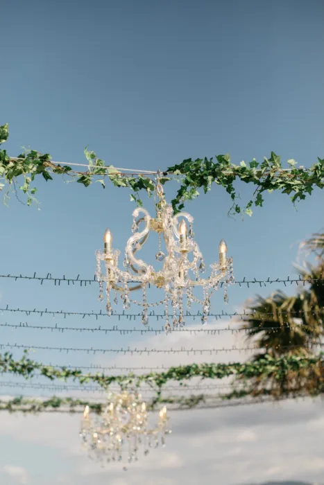 Crystal chandeliers hang above the outdoor reception beneath greenery and open sky.