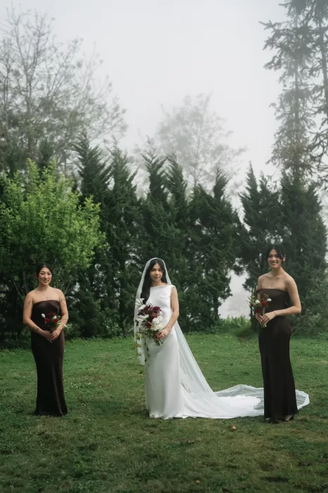 The bride stands with her bridesmaids in dark dresses against a misty green backdrop.