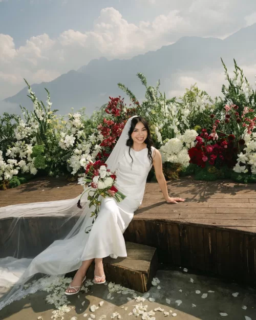The bride sits beside ceremony flowers with her veil falling across the wooden platform.