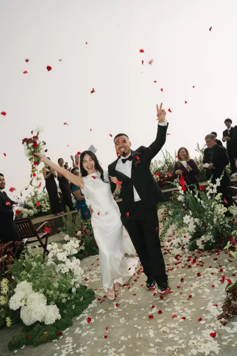 The couple celebrate beneath falling red petals as guests cheer from the ceremony deck.