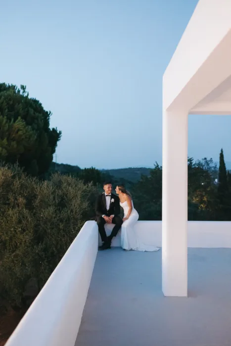 The couple sit quietly on a white terrace as dusk settles over the surrounding hills.