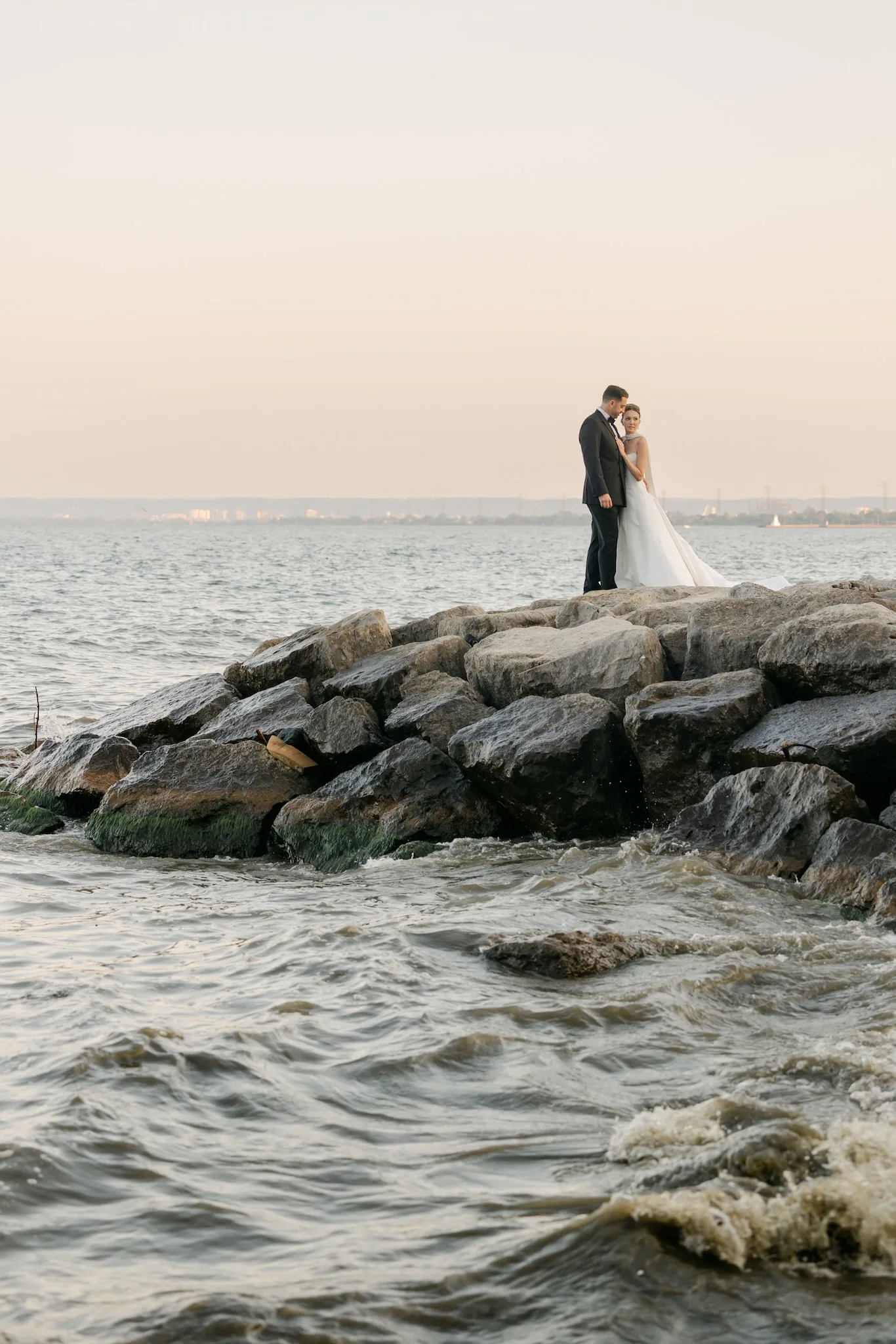 Bride and groom standing on lakeside rocks with soft water behind them.