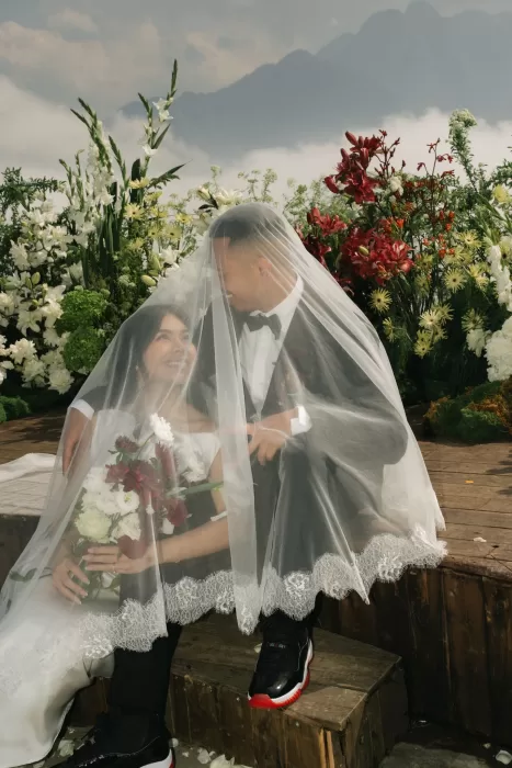 The couple sit beneath the bride’s veil, smiling among mountain florals and soft haze.
