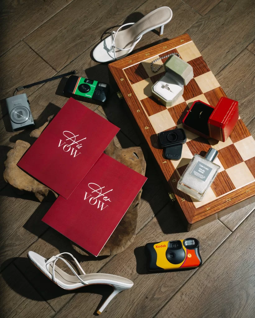 Vow books, rings, heels, cameras and perfume arranged in a sunlit wedding flat lay.