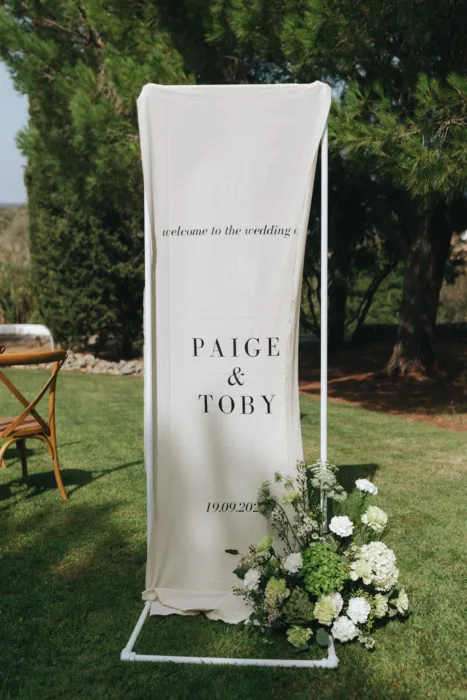 A fabric welcome sign with white florals stands on the lawn before the ceremony.
