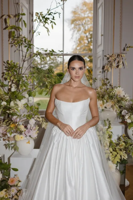 Bride standing among seasonal floral arrangements in front of a tall window.
