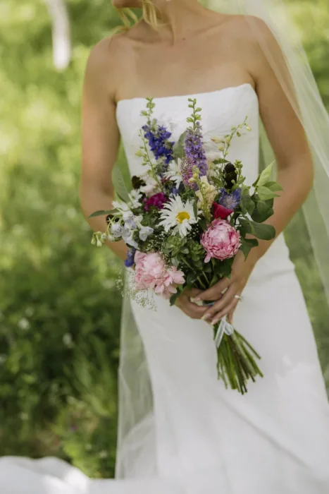 Bride holds a loose bouquet of daisies, peonies and colourful mountain wildflowers.