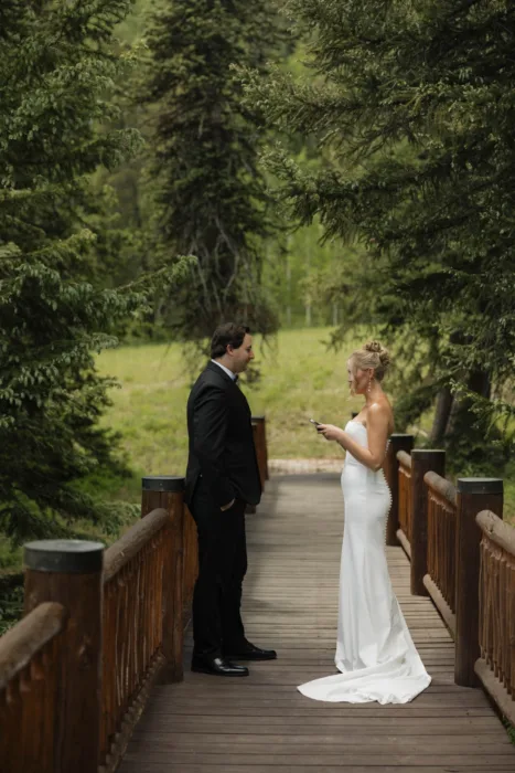 Bride reads to the groom on a wooden bridge surrounded by pine trees.