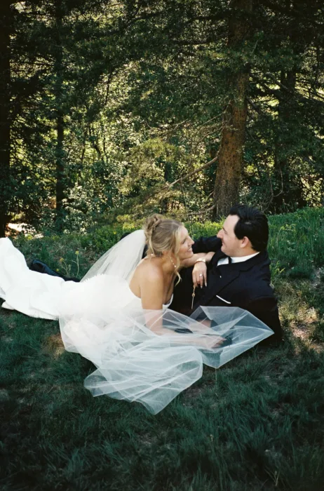 Couple reclines in the forest grass, wrapped in the bride’s veil and dappled light.