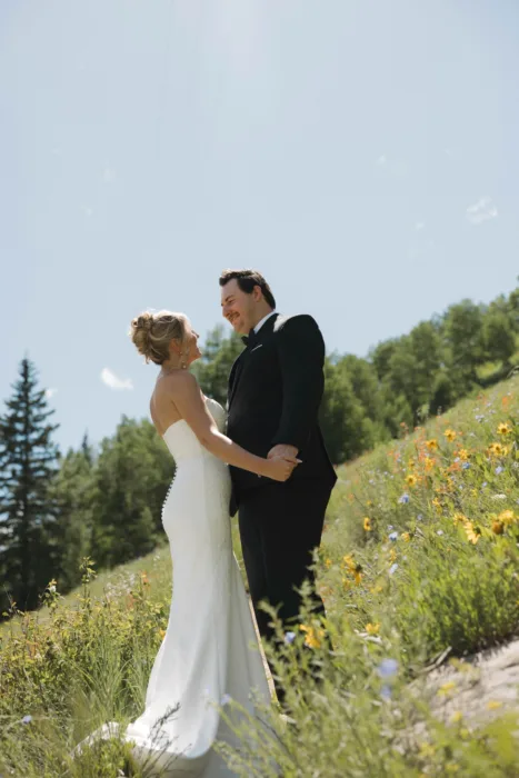 Bride and groom laugh together on a wildflower hillside beneath clear summer sky.