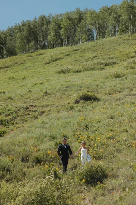 Couple walks through a tall wildflower meadow beneath a clear blue mountain sky.
