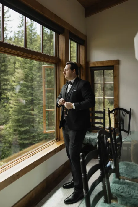 Groom adjusts his black-tie suit beside tall lodge windows overlooking the forest.