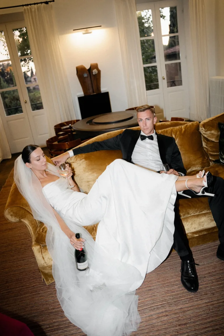 A bride and groom recline on a velvet sofa with champagne in a warm lounge setting.