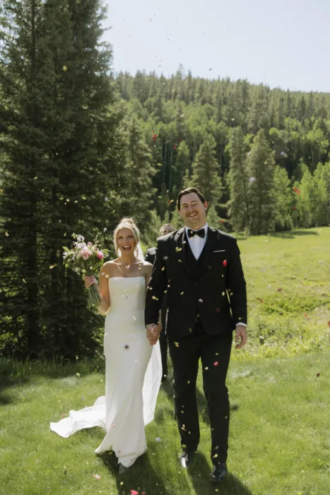 Newlyweds walk through flower confetti in a bright green mountain meadow.