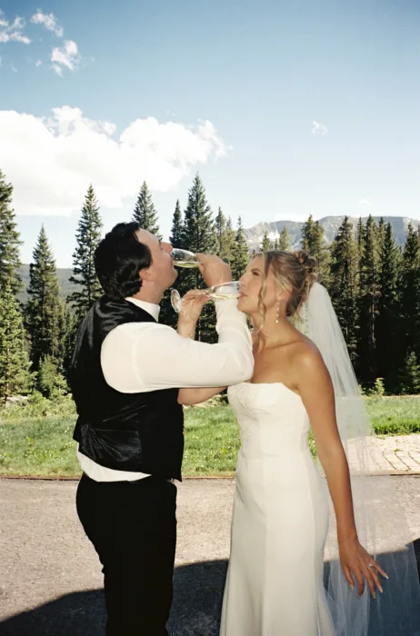 Newlyweds share a champagne toast outdoors with pine trees and mountains behind them.