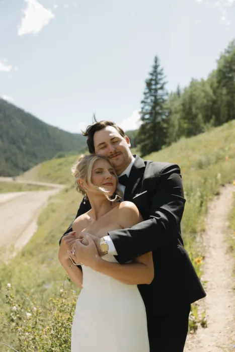 Groom embraces the bride on a quiet mountain road in bright midday sun.