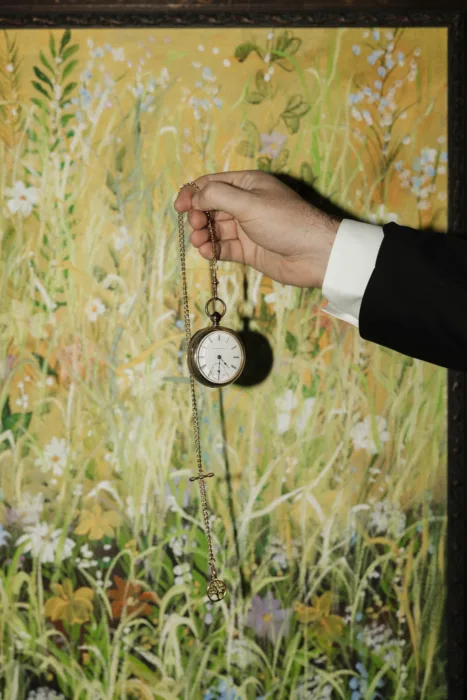 Groom holds a pocket watch against a painted floral backdrop before the wedding.