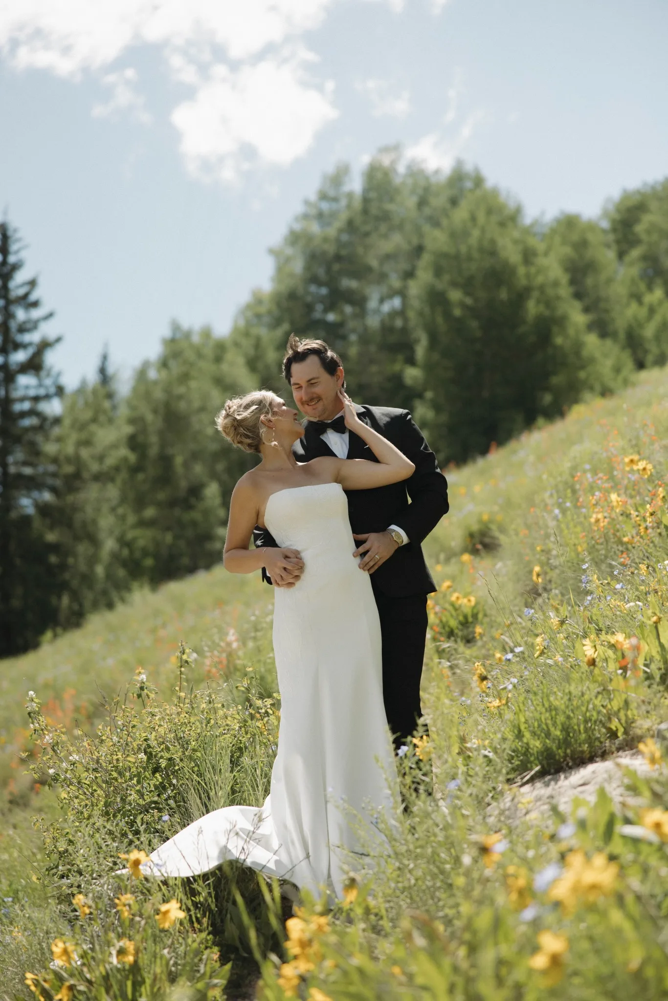 Bride and groom embrace among yellow wildflowers on a sunlit mountain slope.