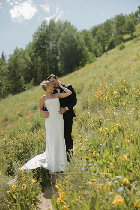 Bride and groom stand close on a narrow trail through bright hillside wildflowers.